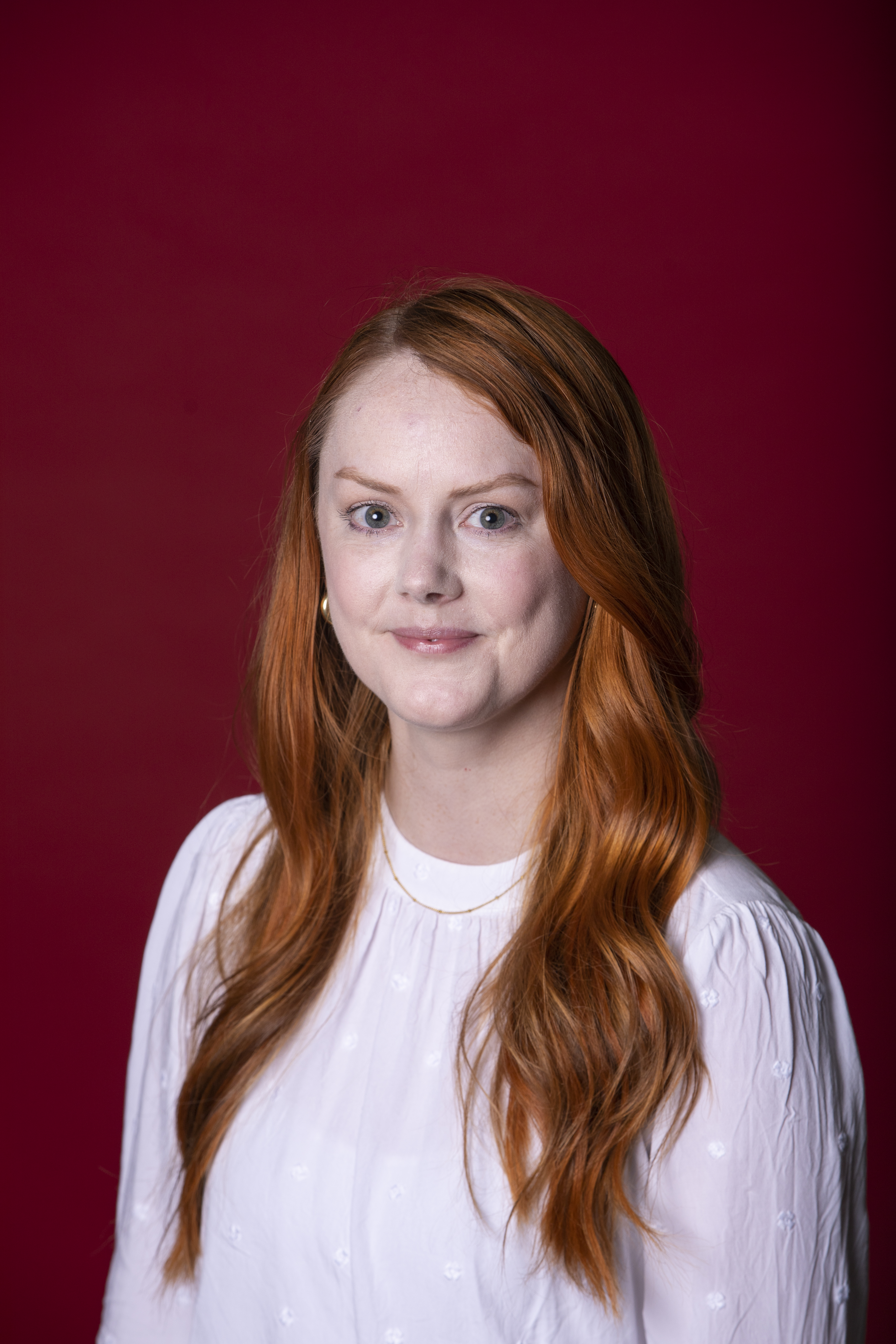 Kara looking at the camera wearing a white blouse and a maroon background.