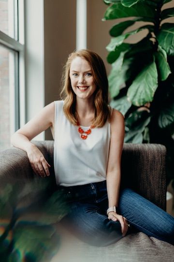 Kara looking at the camera wearing a white blouse. Blurred plants in the background.