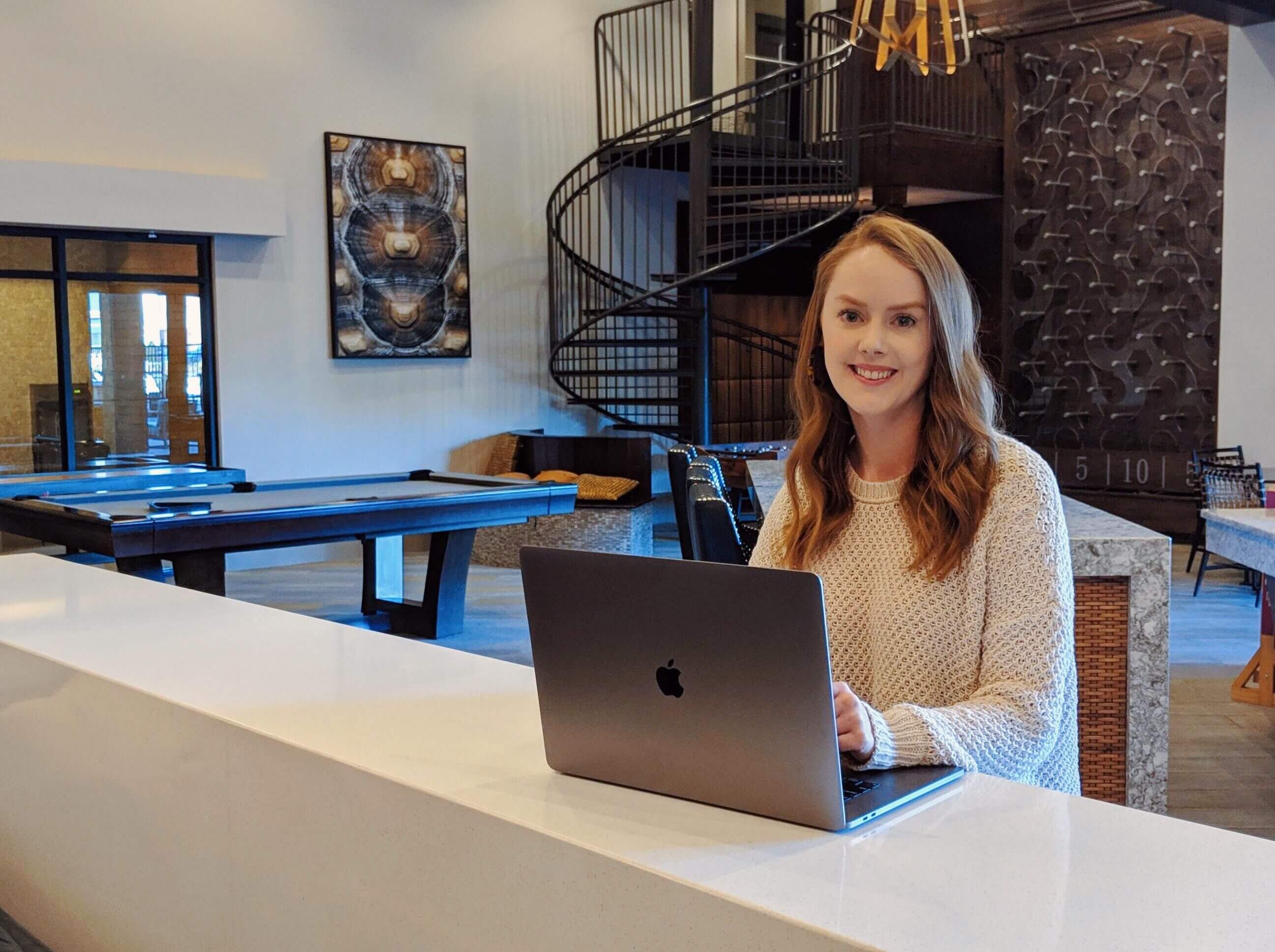 Kara looking at the camera wearing a cream-colored sweater with a laptop in front of her and a recreation room in the background.